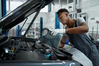 Male worker changes oil in engine, car service. Vehicle repairing garage, man in uniform, automobile station interior