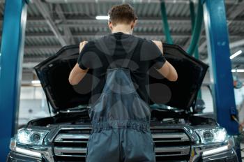Male mechanic at the opened hood, bottom view, car service. Vehicle repairing garage, man in uniform, automobile station interior on background