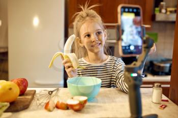 Little girl with banana makes food blog, child blogger. Kid blogging in home studio, social media for young audience, online internet broadcast, creative hobby