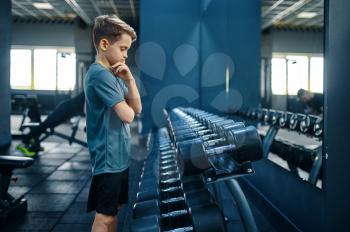 Boy tries to take heavy dumbbell in gym. Youngster on training in sport club, healthcare and healthy lifestyle, schoolboy on workout