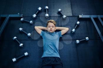 Boy lying on mat among many dumbbells in gym. Youngster on training in sport club, healthcare and healthy lifestyle, schoolboy on workout, sportive youth