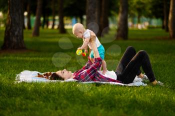Mother and her little baby lying on grass in summer park. Mom with male kid on lawn, picnic with child on plaid in the forest, family happiness