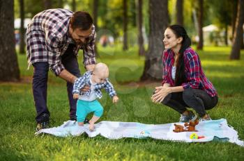 Happy parents and little baby play on grass in summer park. Mom and dad with male kid on lawn, picnic with child on plaid in the forest, family happiness