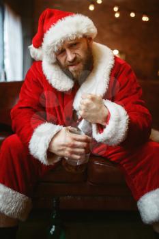Bad Santa claus celebrate christmas with bottle of alcohol on couch. Unhealthy lifestyle, bearded man in holiday costume