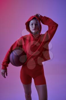 Sexy female athlete in red hoodie poses with ball in studio, neon background. Fitness sportswoman at the photo shoot, sport concept, active lifestyle motivation