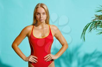 Sportive woman in red swimsuit poses on cyan background in studio. Girl in swimwear ready to take a tan. Model with slim body in swimming underwear, seductive female swimmer