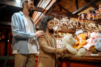 Love couple wins soft toy in amusement park souvenir shop. Man and woman relax outdoors, fairground on background. Family leisures on carousels, entertainment theme