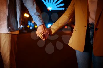Love couple holding hands in night amusement park. Man and woman relax outdoors, ferris wheel with lights on background. Family leisures on carousels, entertainment theme