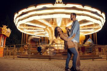 Love couple dancing at the carousel in night amusement park. Man and woman relax outdoors, roundabout attraction with lights on background. Family leisures in summertime, entertainment theme