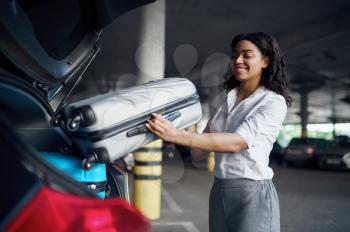 Young woman puts suitcases into the car on parking. Female traveler with luggage in vehicle park lot, passenger with many bags. Girl with baggage near automobile