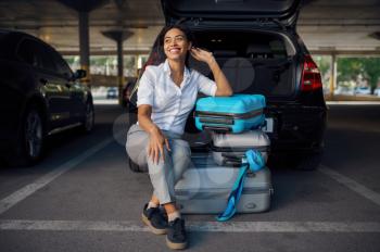 Happy woman sitting on suitcases in car parking. Female traveler with luggage in vehicle park lot, passenger with bag. Girl with baggage near automobile