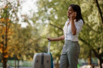 Young woman with suitcase talking by phone in park. Female traveler with luggage leisures outdoors, passenger with bag resting in nature. Girl with baggage relax on city alley