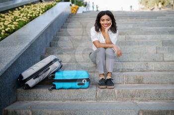 Young woman with suitcase sitting on the stairs in summer park. Female traveler with luggage leisures outdoors, passenger with bag resting in nature. Girl with baggage relax on city alley