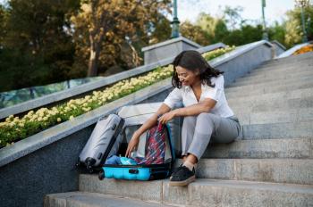 Young woman opens suitcase on the stairs in summer park. Female traveler with luggage leisures outdoors, passenger with bag resting in nature. Girl with baggage relax on city alley