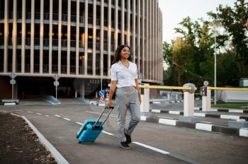 Young woman with suitcase at the entrance to car parking. Female traveler with luggage near vehicle park lot, passenger with bag. Girl with baggage on city street