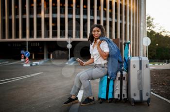 Young woman sitting on suitcase at the entrance to car parking. Female traveler with luggage near vehicle park lot, passenger with bag. Girl with baggage on city street