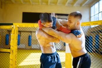 Male MMA fighter performs painful hold to his opponent in a cage in gym. Muscular men on ring, combat workout, martial arts training, competition or sparring