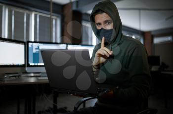 Male internet hacker in hood works on laptop in dark office. Illegal web programmer at workplace, criminal occupation. Data hacking, cyber security