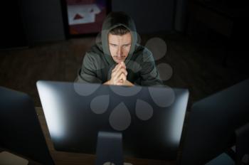 Serious male internet hacker in hood works on computer, front view. Illegal web programmer at workplace, criminal occupation. Data hacking, cyber security
