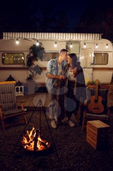 Young love couple kissing by the campfire, picnic at camping in the forest. Summer adventure on rv, camping-car on background. Man and woman leisures by the bonfire, travelling with trailer