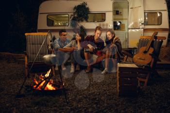 Friends sitting by the campfire in the night, picnic at camping in the forest. Youth having summer adventure on rv, camping-car on background. Two couples leisures, travelling with trailer