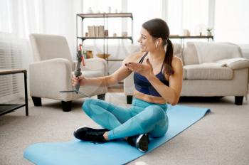 Slim woman sits on mat, online fit training with phone. Female person in sportswear, internet sport workout, room interior on background
