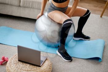Woman sitting on ball, online pilates training at the laptop. Female person in sportswear, internet sport workout, room interior on background