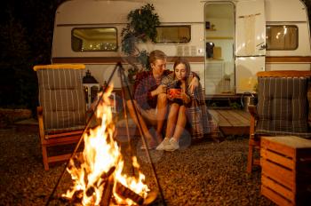 Young love couple hugging by the campfire, picnic at camping in the forest. Summer adventure on rv, camping-car on background. Man and woman leisures by the bonfire, travelling with trailer