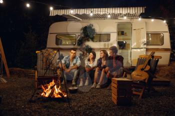 Friends sitting by the campfire in the night, picnic at camping in the forest. Youth having summer adventure on rv, camping-car on background. Two couples leisures, travelling with trailer