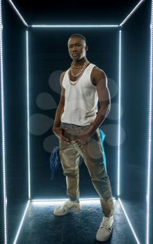 Serious rapper in gold jewelry poses in illuminated cube, dark background. Hip-hop performer, rap singer, break-dance performing, entertainment lifestyle, breakdancer