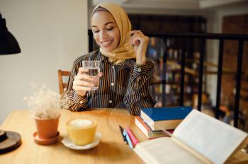 Arab girl in hijab holds glass of water, university cafe interior on background. Muslim woman with books sitting in library. Religion and education