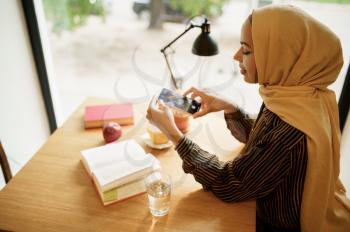 Arab girl makes photo of cup with coffee in university cafe, top view. Muslim woman with books sitting in library. Religion and education