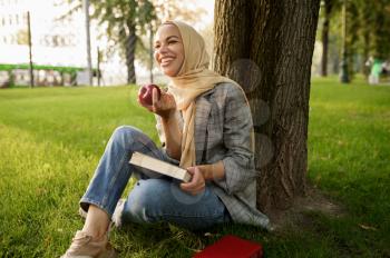 Smiling arab girl in hijab holds apple and textbook in summer park. Muslim woman with books resting on the lawn. Religion and education