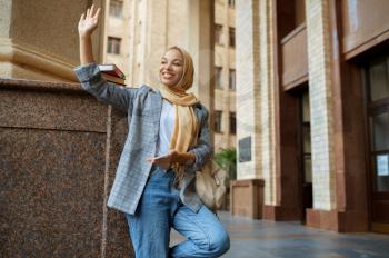 Arab female student in hijab makes selfie at the university entrance. Muslim woman with books poses in front of a college. Religion and education