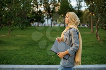 Arab female student with laptop walking in summer park. Muslim woman resting on the walking path. Religion and education