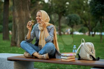 Arab female student sitting on the bench in summer park. Muslim woman resting on the walking path. Religion and education