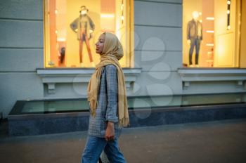 Arab girl in hijab looking on showcase of fashion store in downtown. Muslim woman walking on street. Female student in big city