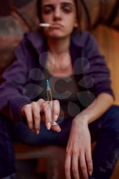 Drug addict female person with cigarette holds syringe in high, shebang interior on background. Narcotic addiction problem, eternal depression of junky people