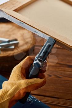 Male hand in glove holds stapler, closeup, wooden background. Professional instrument, carpenter equipment, woodworker tools