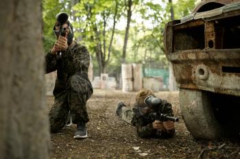 Two male warriors in camouflages and masks aimimg with paintball guns. Extreme sport with pneumatic weapon and paint bullets or markers, military team game outdoors, combat tactics