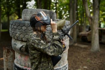 Female paintball player with gun poses on playground in the forest. Extreme sport with pneumatic weapon and paint bullets or markers, military team game outdoors, combat tactics