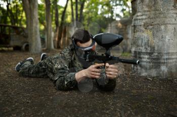Male paintball player with gun shoots lying down on the ground, playground in the forest on background. Extreme sport with pneumatic weapon and paint bullets or markers, military team game outdoors