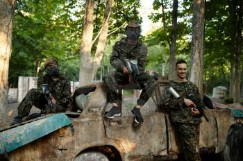 Paintball team, friends poses at rusty car on playground in the forest. Extreme sport with pneumatic weapon and paint bullets or markers, military game outdoors, combat tactics