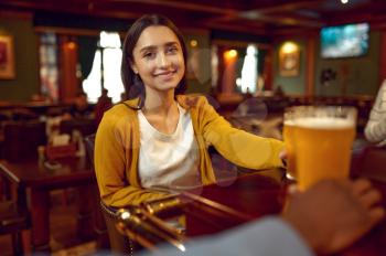 Smiling girl holds glass of beer at the counter in bar. Group of people relax in pub, night lifestyle, friends celebrate event in restaurant