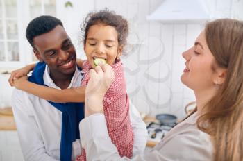 Cheerful family, nice breakfast on the kitchen. Mother, father and their funny daughter cooking in the morning, good relationship. Little child tastes a cucumber