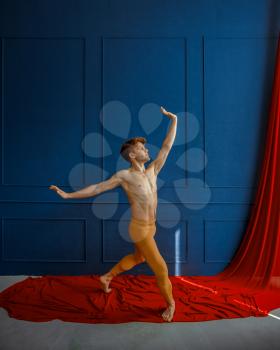 Male ballet dancer, performing in action, dancing studio, blue wall and red cloth on background. Performer with muscular body, grace and elegance of movements