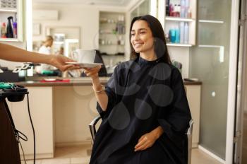 Hairdresser gives cup of coffee to female customer, hairdressing salon. Stylist and client in hairsalon. Beauty business, professional service