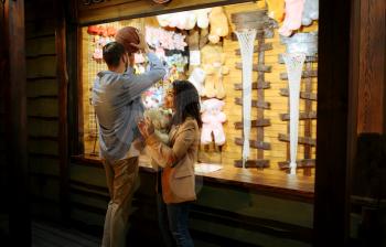 Love couple play in shooting gallery, night amusement park. Man and woman relax outdoors, fairground on background. Family leisures on carousels, entertainment theme