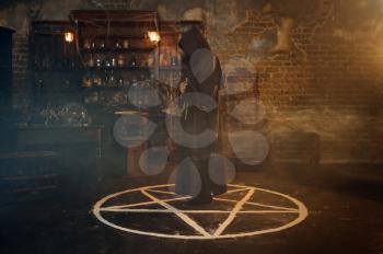 Male exorcist in black hood standing in the magic circle. Exorcism, mystery paranormal ritual, dark religion, night horror, potions on shelf on background