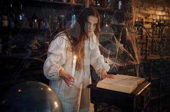 Young demonic woman with candle reads book of spells, demons casting out. Exorcism, mystery paranormal ritual, dark religion, night horror, potions on shelf on background